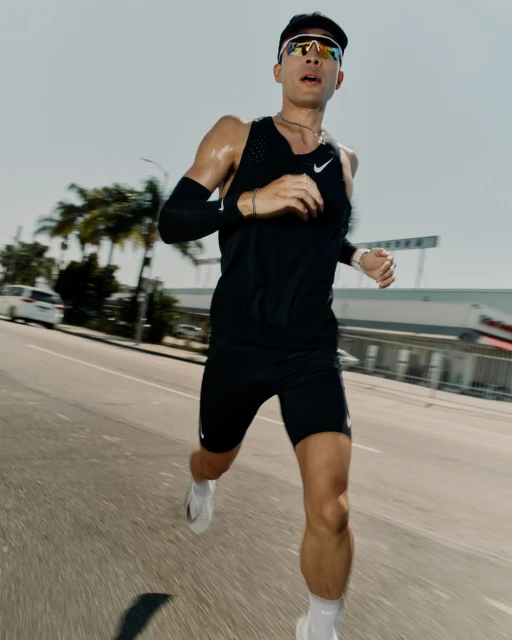A male runner in athletic wear and sunglasses is captured in motion on a city street, with buildings and palm trees in the background.