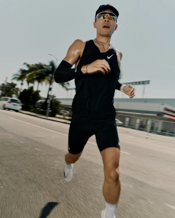A male runner in athletic wear and sunglasses is captured in motion on a city street, with buildings and palm trees in the background.