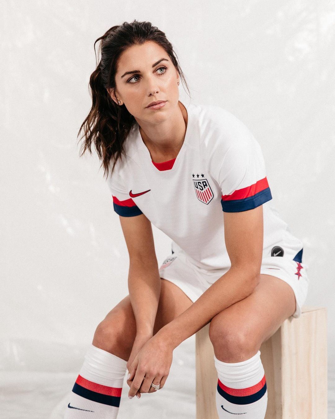 Portrait of a woman in a soccer uniform sitting on a stool, looking off to the side, against a white backdrop in a photo studio.