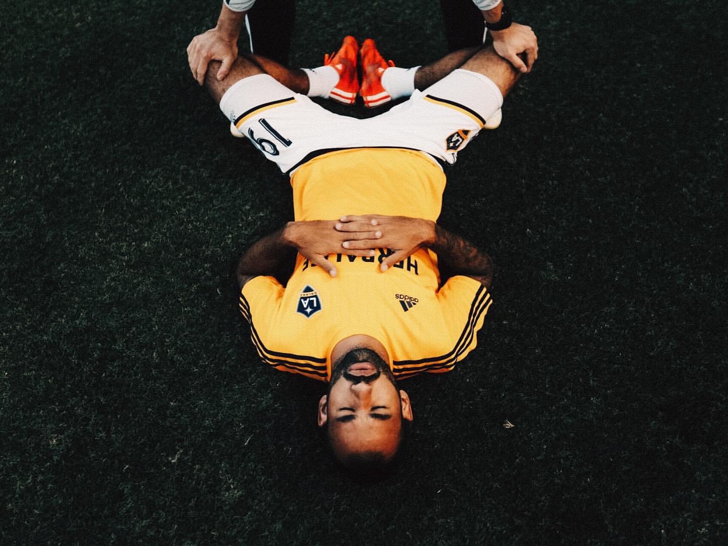 Overhead shot of a soccer player lying on a grassy field with his legs being stretched by two people, wearing a yellow jersey.