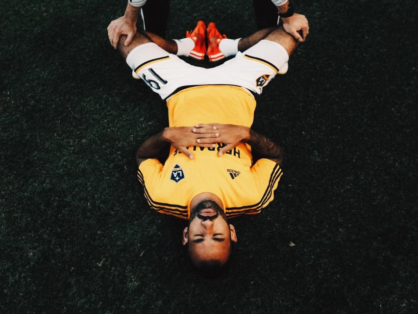 Overhead shot of a soccer player lying on a grassy field with his legs being stretched by two people, wearing a yellow jersey.