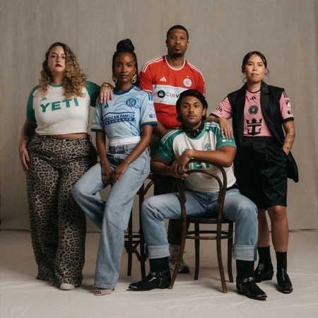 Group of five diverse adults wearing soccer jerseys pose in a studio, showcasing team apparel against a neutral backdrop.