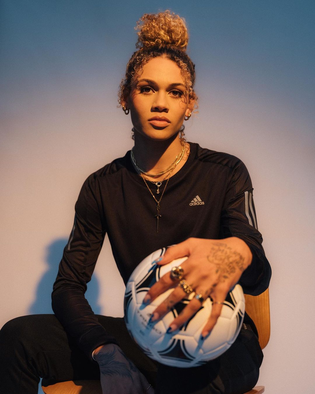A mixed-race adult in sport clothing sits while holding a soccer ball, wearing a black long-sleeved shirt, multiple rings, and a necklace.
