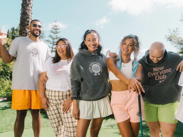 Group of five people outdoors, wearing t-shirts and hoodies with the text 'New Everyone Club' against a sunny backdrop.