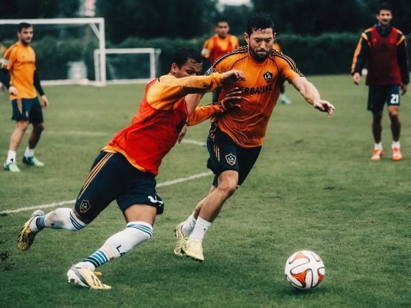 Two adult male soccer players in action on a green field with a soccer ball during a daytime game, surrounded by other players.