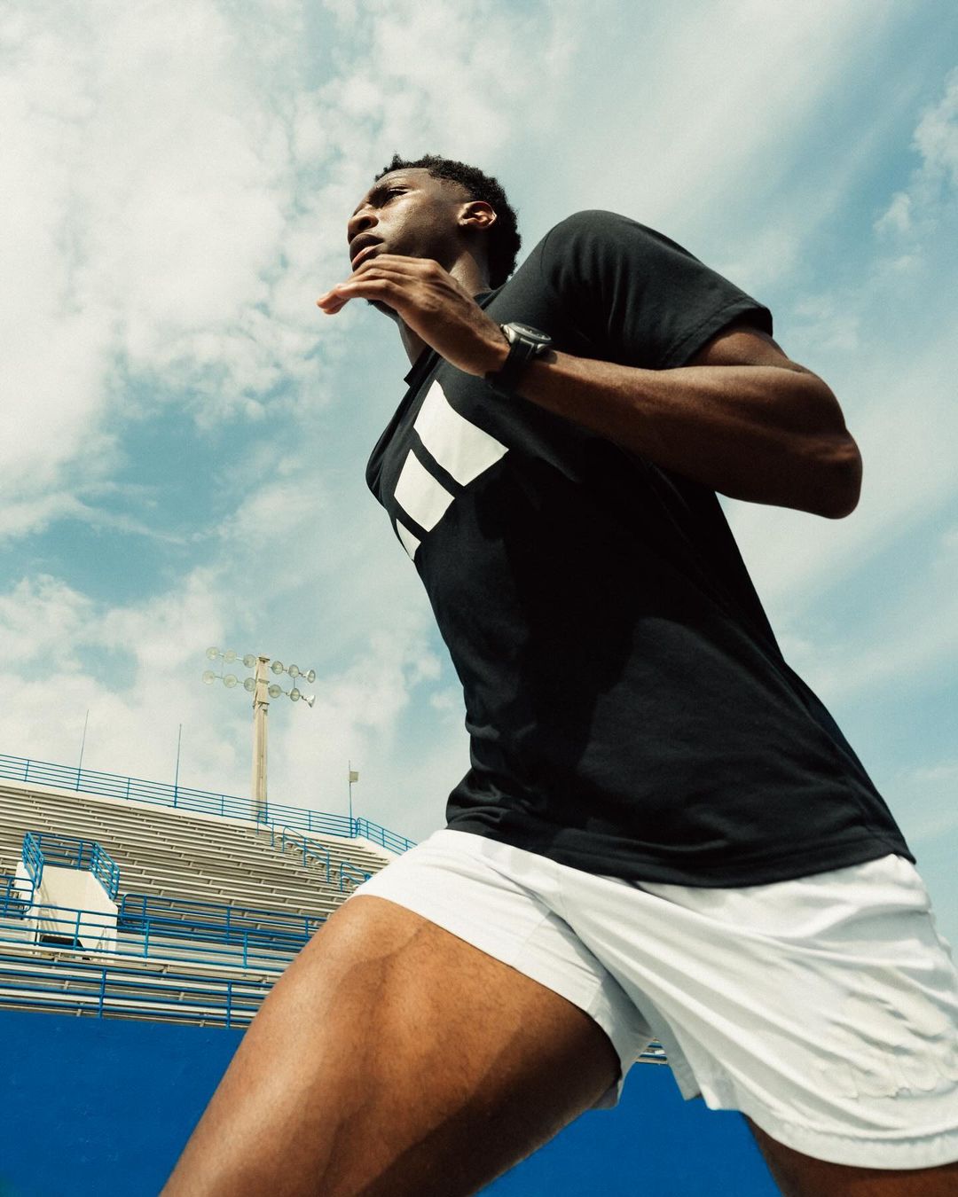 Athletic Black male runner in black t-shirt and white shorts. Running against blue sky in outdoor stadium. Low angle, full shot.