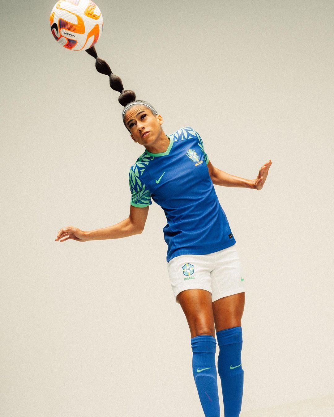 A woman in a blue soccer jersey balancing a soccer ball on her head, in a light, neutral studio setting. She is mid-motion with outstretched arms.