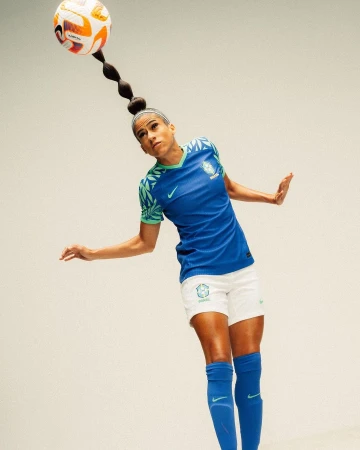 A woman in a blue soccer jersey balancing a soccer ball on her head, in a light, neutral studio setting. She is mid-motion with outstretched arms.