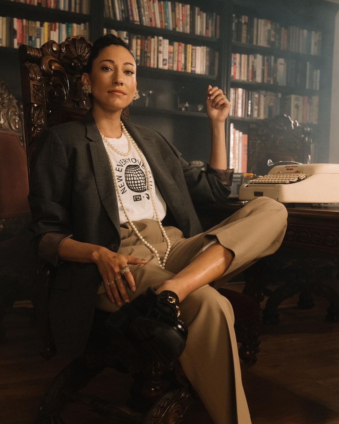 Indoor portrait of an adult woman sitting in a wooden armchair wearing a white t-shirt, blazer, and beige pants in front of a bookshelf.
