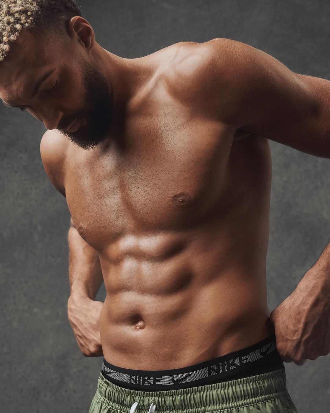 Athletic adult male showing muscular physique in a studio shot, wearing green athletic shorts with the Nike logo on the waistband.
