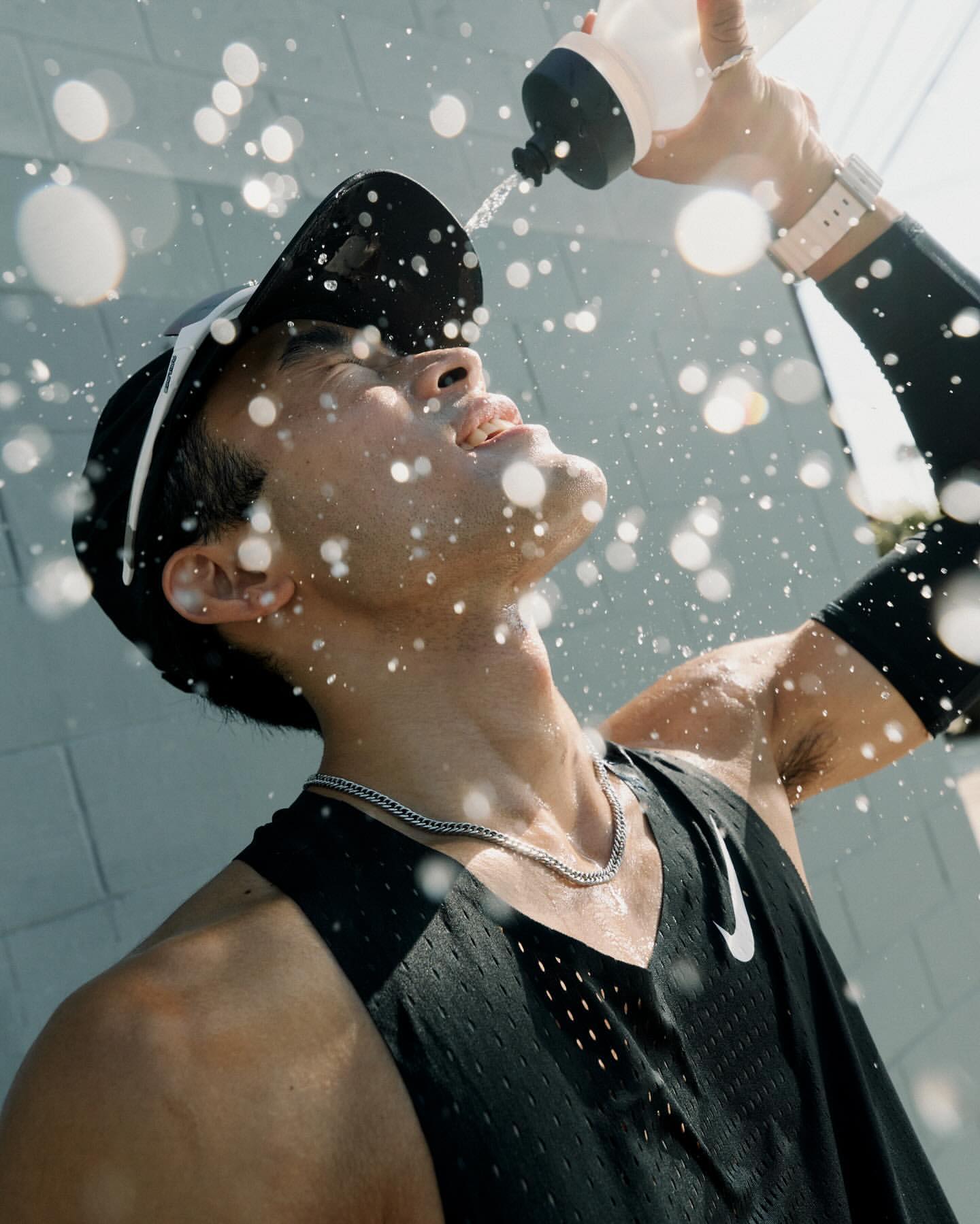 Athletic man in black sports gear pouring water over his head outdoors on a sunny day, water droplets visible in the bright light.