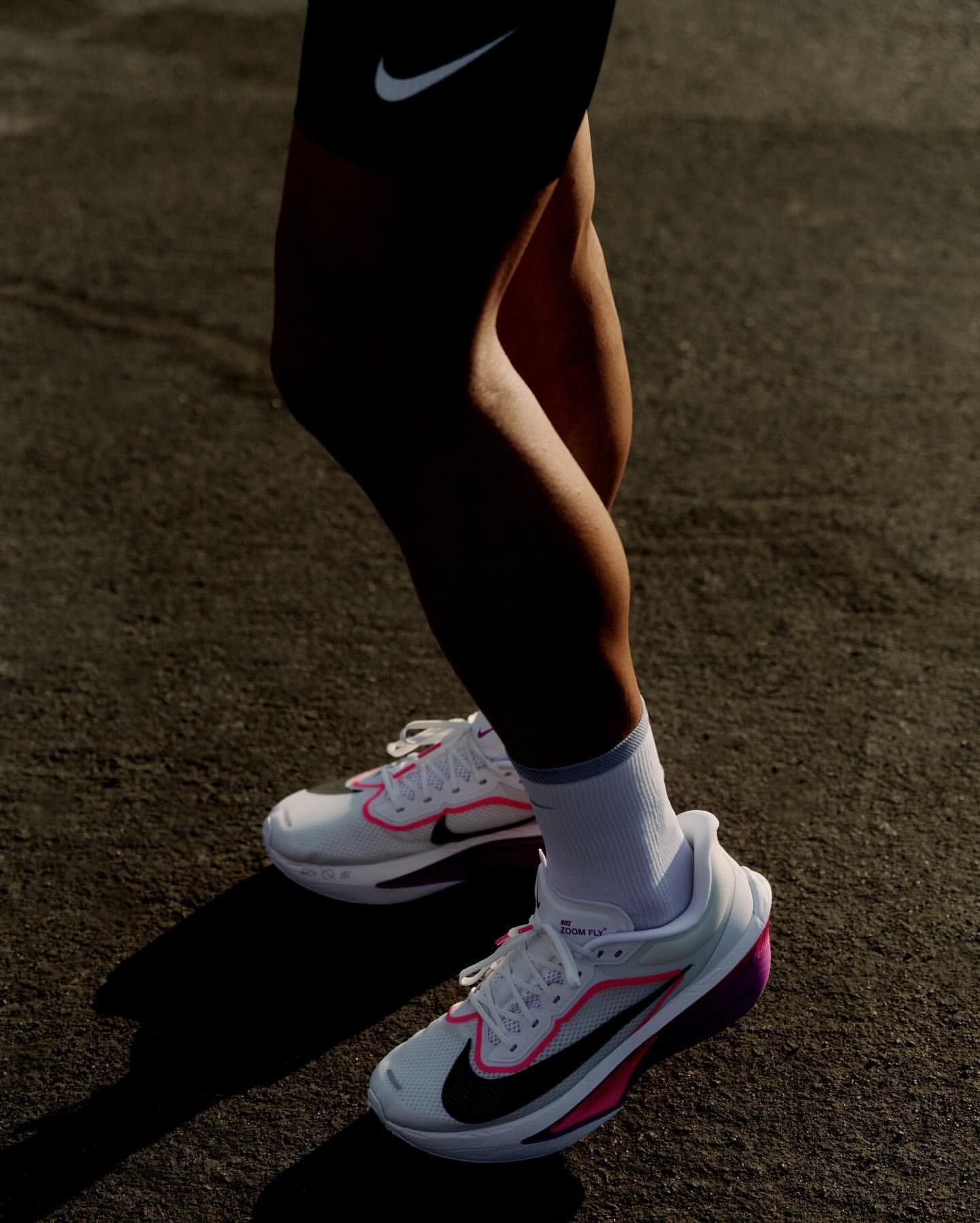 A pair of Nike running shoes on asphalt. The shoes are white with a black Nike swoosh and pink trim. Legs and feet are visible.