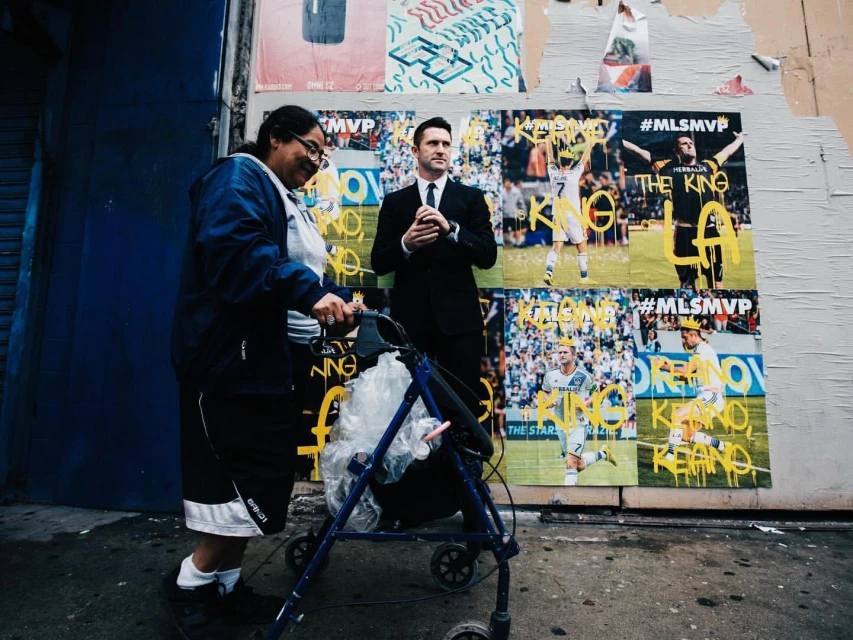 Woman with walker and man in suit walk past posters of soccer players on a city street. Outdoor scene with urban grit and dark shadows.