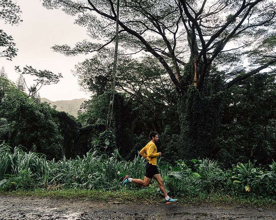 Man running on a dirt path surrounded by lush green vegetation and trees, wearing a yellow long-sleeve shirt and black shorts.