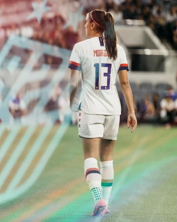 A woman in a white soccer jersey with the number 13 on her back is walking on a green field with a blurred crowd in the background.