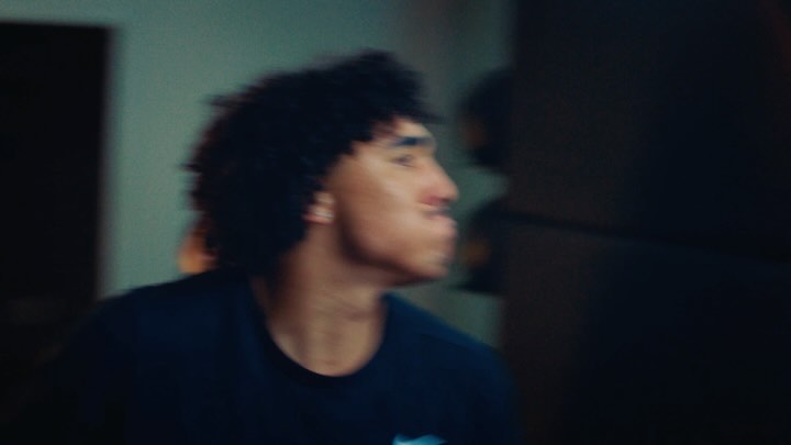 Close-up shot of a young adult with curly hair wearing a dark t-shirt indoors, against a blurry teal-toned wall and a dark background.