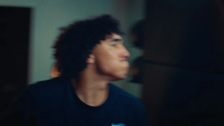 Close-up shot of a young adult with curly hair wearing a dark t-shirt indoors, against a blurry teal-toned wall and a dark background.