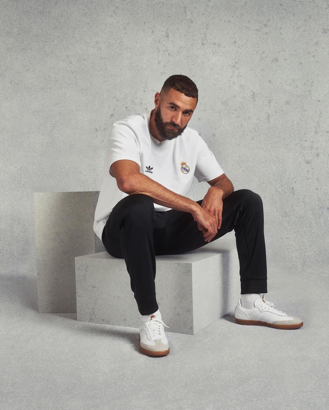 Man in a white t-shirt with the Real Madrid logo sits on a white cube against a textured gray background in a studio shot.