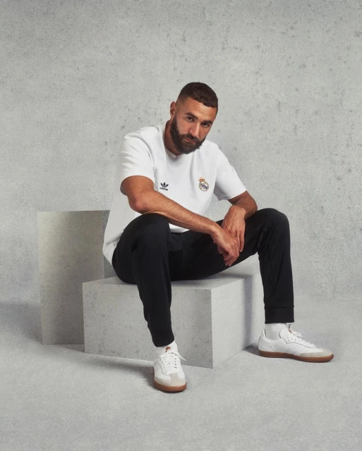 Man in a white t-shirt with the Real Madrid logo sits on a white cube against a textured gray background in a studio shot.