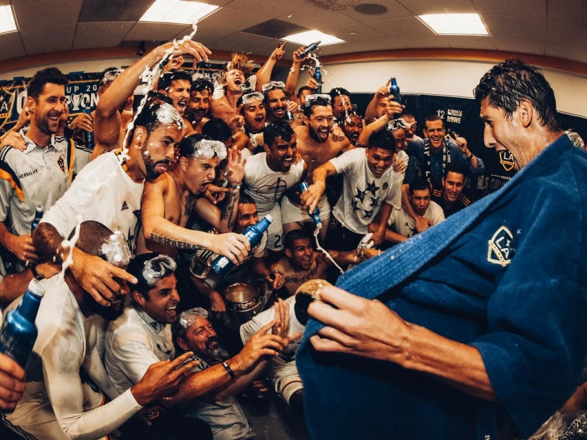 Soccer team celebrating a championship win in a locker room, covered in foam and holding beer bottles, with one player in a robe.