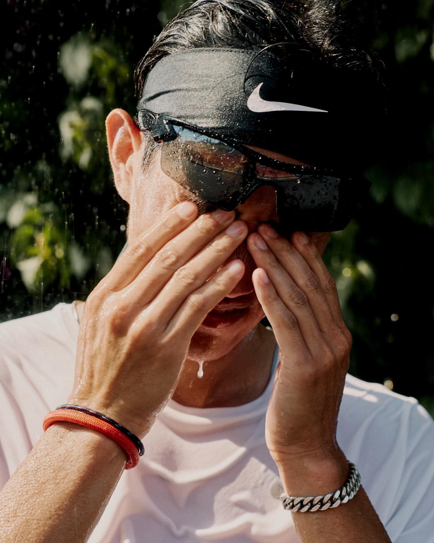 Sweaty man in athletic wear and sunglasses wipes his face in an outdoor setting with a blurred green leafy background and water droplets.
