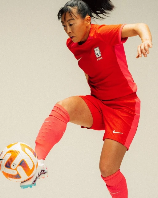 Adult female soccer player in red Korean national team uniform kicking a white soccer ball in an indoor photo studio setting.