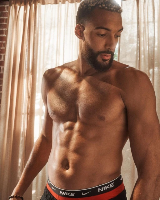 Adult male with beard, posing shirtless indoors with a brick wall and curtain background in a medium shot; Nike branded underwear.