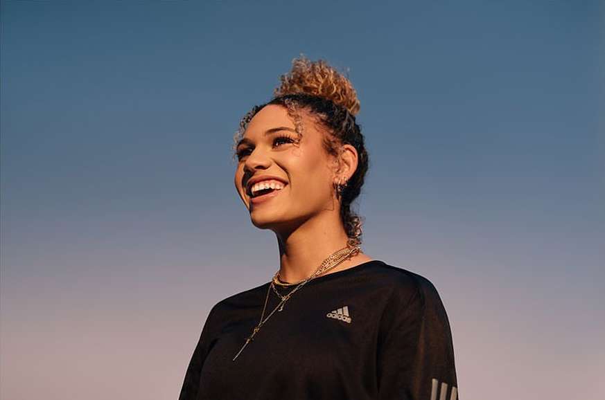 A smiling adult woman with curly hair in a bun, wearing a black Adidas long-sleeve shirt and gold necklaces, set against a blue gradient sky.