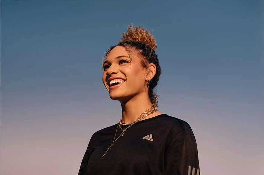 A smiling adult woman with curly hair in a bun, wearing a black Adidas long-sleeve shirt and gold necklaces, set against a blue gradient sky.