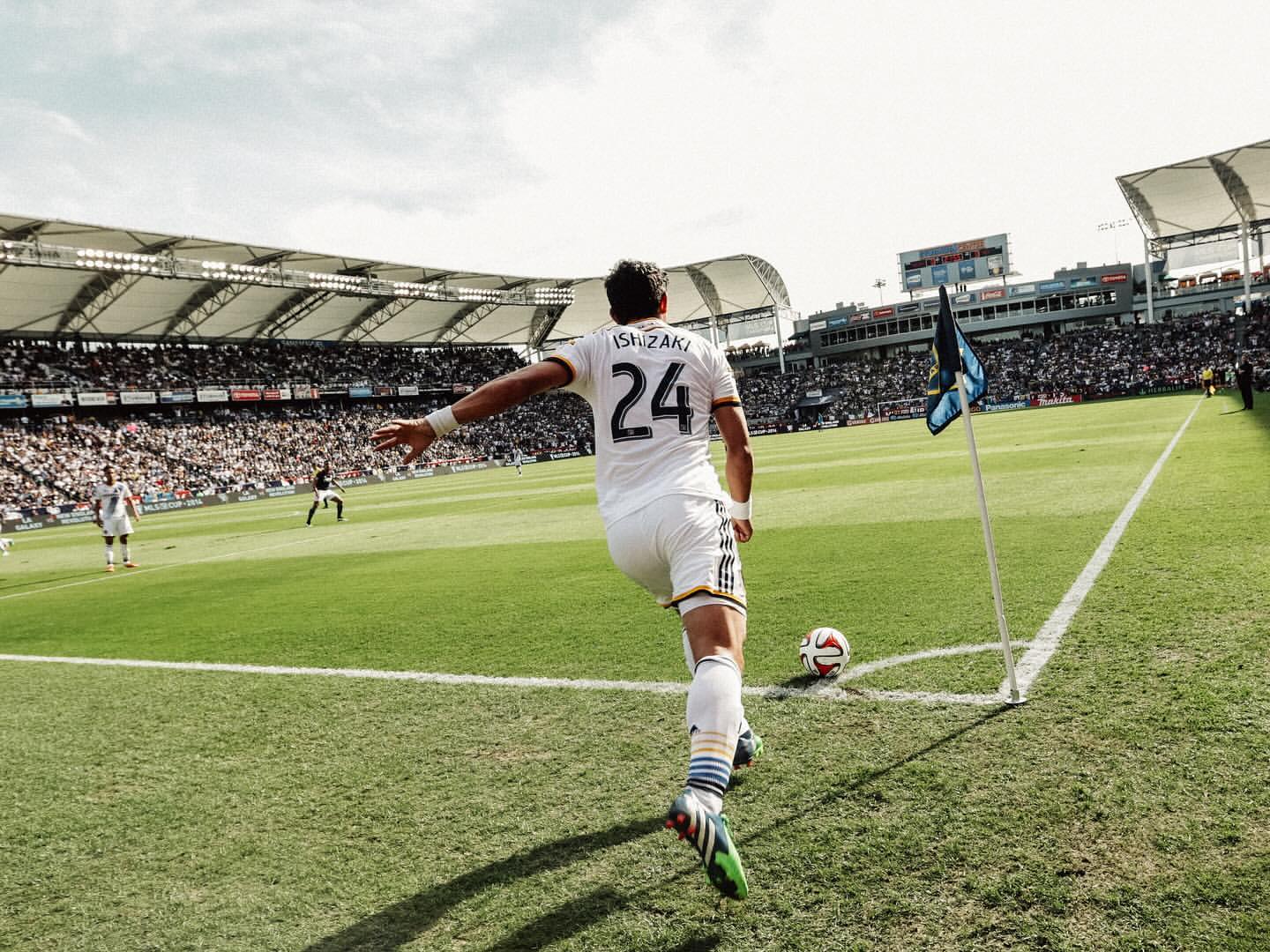 A soccer player in a white uniform is about to kick a soccer ball at the corner of a vibrant green field in a stadium filled with fans.