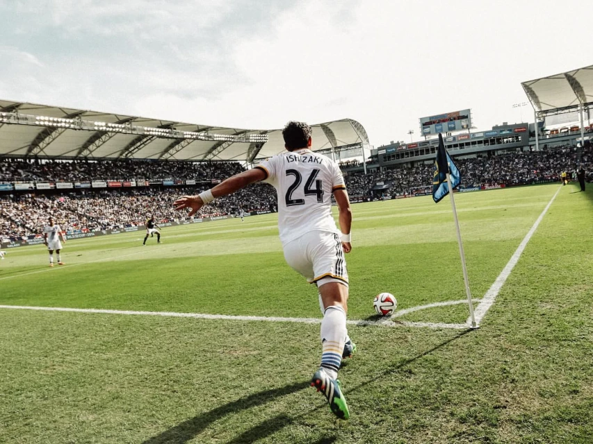 A soccer player in a white uniform is about to kick a soccer ball at the corner of a vibrant green field in a stadium filled with fans.