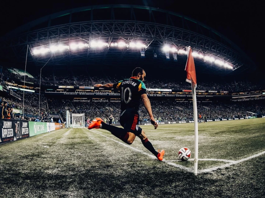 Soccer player kicks ball on the field in a stadium at night; a corner flag stands nearby under bright stadium lights.