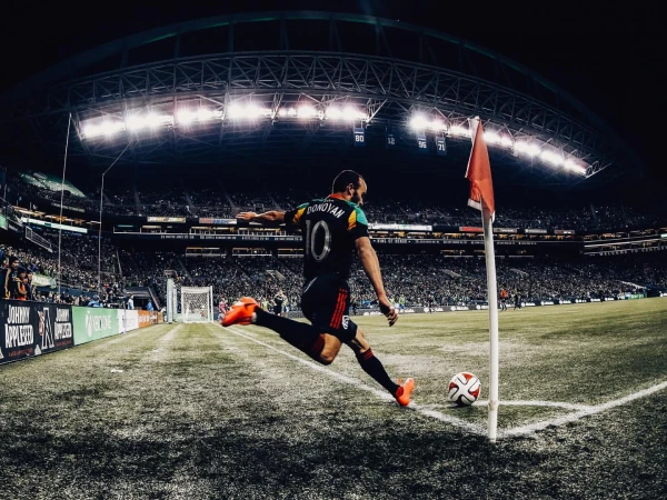 Soccer player kicks ball on the field in a stadium at night; a corner flag stands nearby under bright stadium lights.