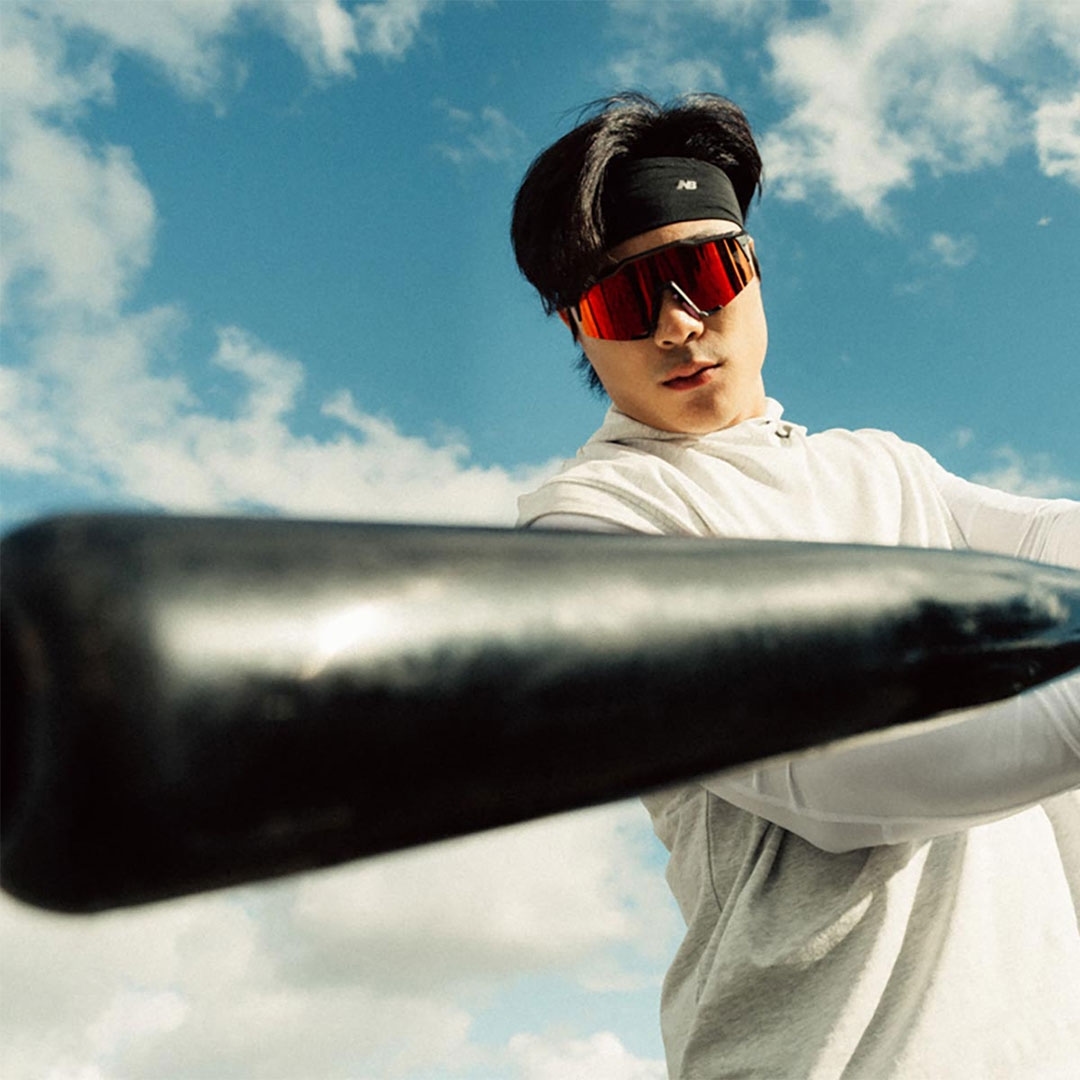 Asian man wearing red sports glasses and a black headband holding a bat outside against a blue sky and white clouds.