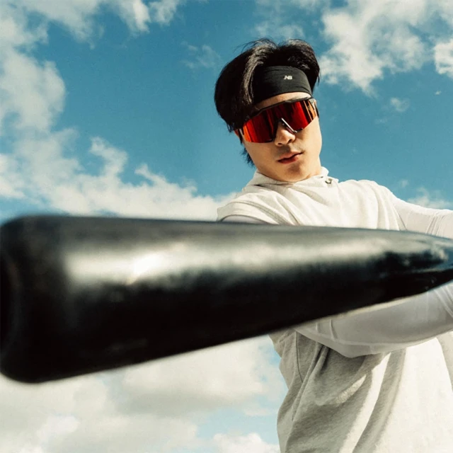 Asian man wearing red sports glasses and a black headband holding a bat outside against a blue sky and white clouds.
