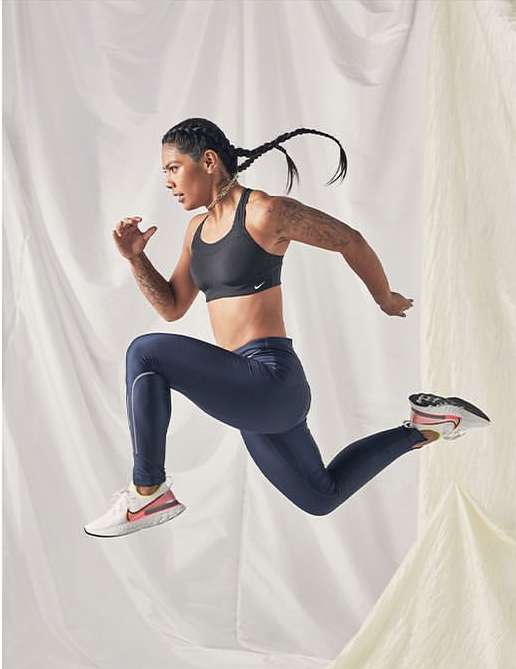 Athletic woman in black sports bra and blue leggings jumps mid-air against a white fabric background in a fitness-themed studio shot.