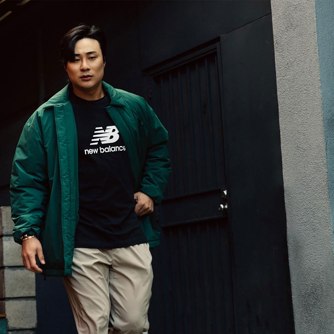 Asian male model in green jacket and khakis poses in front of a black metal door with silver hardware, urban background, natural light.