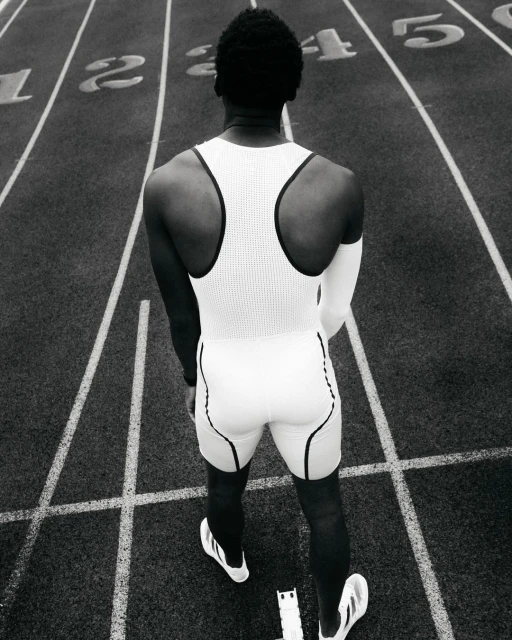 Back view of a Black male athlete in a white uniform on a running track, focused on the start line with lane numbers, monochrome.