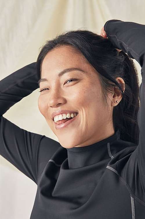 Portrait of a smiling Asian woman with dark hair, wearing a long-sleeved black shirt and putting her hair up, set against a neutral background.