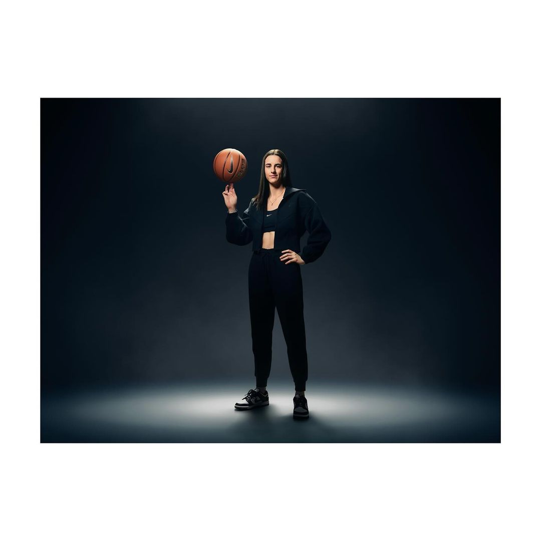 A woman in dark athletic wear spins a basketball on her finger in a full shot against a dark grey background with dramatic lighting.