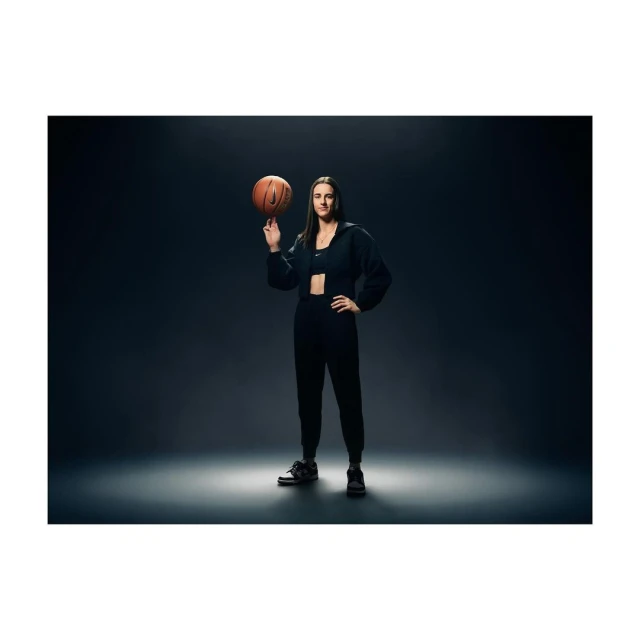 A woman in dark athletic wear spins a basketball on her finger in a full shot against a dark grey background with dramatic lighting.