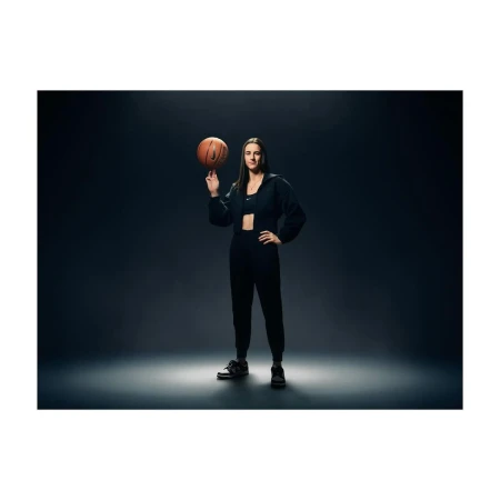 A woman in dark athletic wear spins a basketball on her finger in a full shot against a dark grey background with dramatic lighting.