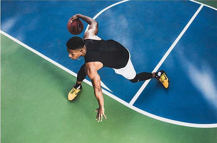 Overhead view of a male basketball player in a black shirt and white shorts dribbling a basketball on a blue and green court.