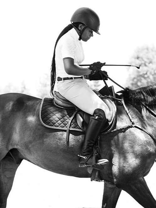 A person wearing a helmet riding a horse, holding a riding crop, wearing riding boots and breeches, in a black and white outdoor shot.
