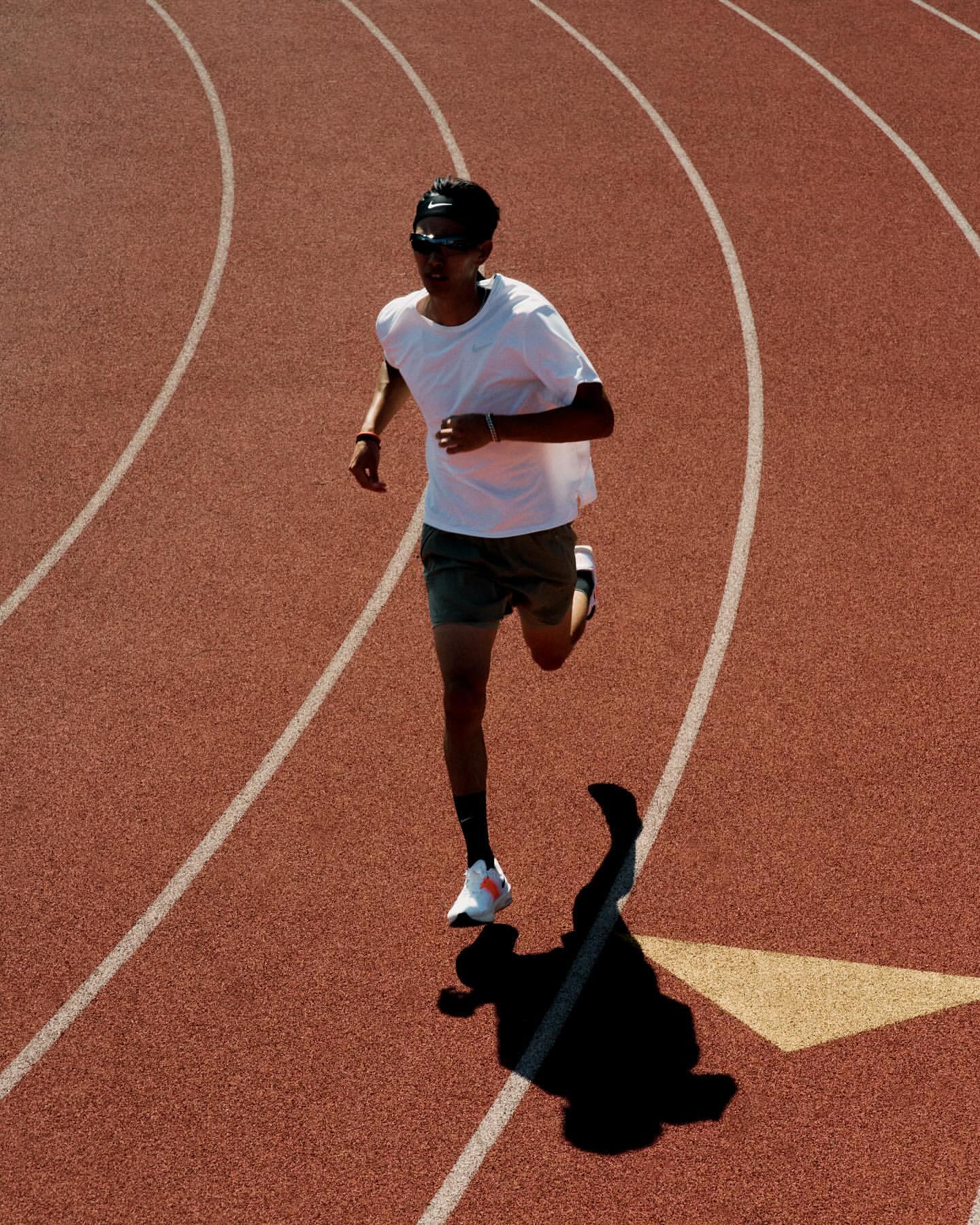 Overhead shot of an adult male wearing sunglasses, a white shirt, and dark shorts jogging on a red track with white lines in sunlight.