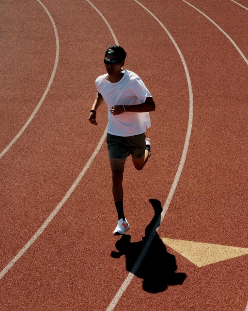 Overhead shot of an adult male wearing sunglasses, a white shirt, and dark shorts jogging on a red track with white lines in sunlight.