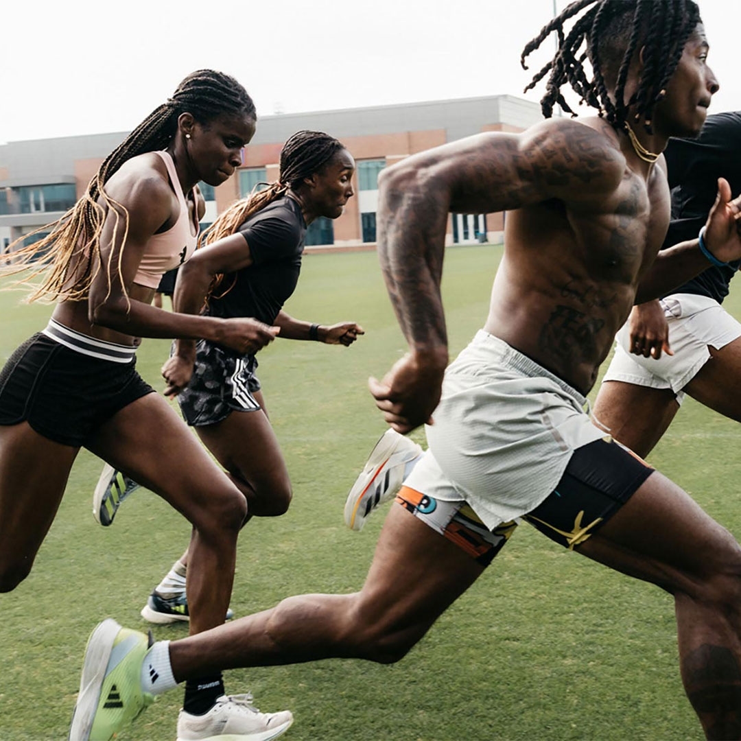 Athletes in running shorts and sports tops sprint on a green field on a cloudy day. Building visible in the background.