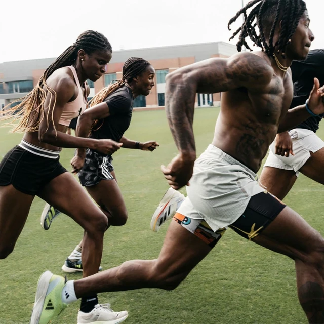 Athletes in running shorts and sports tops sprint on a green field on a cloudy day. Building visible in the background.