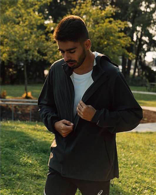 Adult male in sportswear zipping up a black hooded jacket in outdoor park setting with trees and lawn in soft, warm daylight.