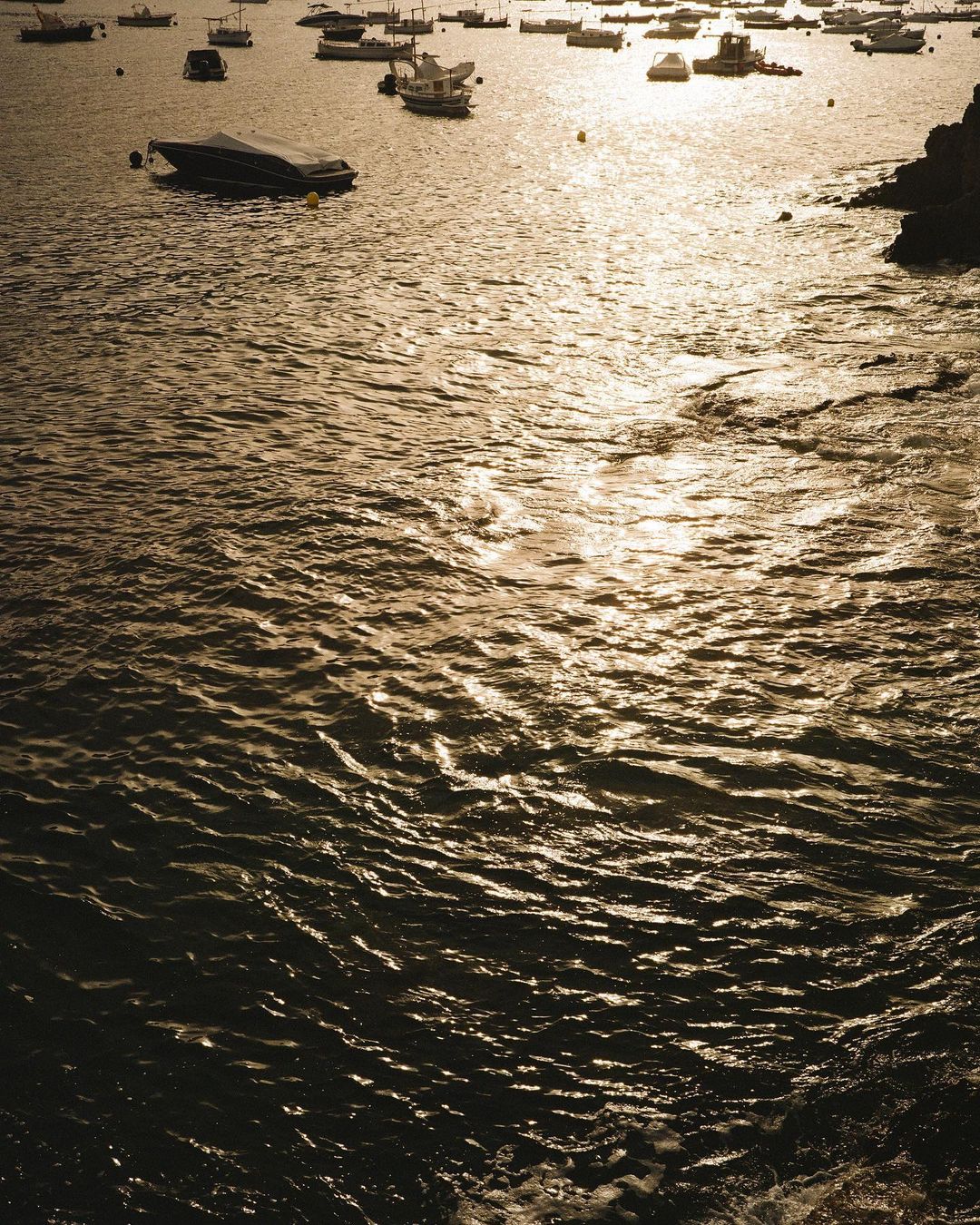 Golden sunlight glistens on the rippling ocean water near a rocky coastline, with several boats visible on the horizon at dusk.
